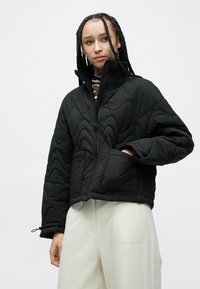 Czarna pikowana kurtka z wysokim kołnierzem, zamkiem błyskawicznym i przednią kieszenią. Wykonana z teksturowanej tkaniny, z elastycznymi mankietami. Noszona na jasnych spodniach.