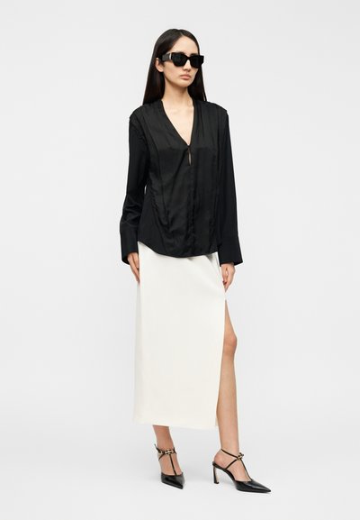 Μαύρη σατέν μπλούζα μακρυμάνικη με V-λαιμόκοψη και πιετέ λεπτομέρειες, συνδυασμένη με λευκή midi φούστα με ψηλό σκίσιμο και μαύρες μυτερές γόβες.