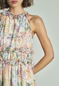 Abito floreale con colori pastello, scollatura e orlo arricciati, spalline con cordoncino e vita arricciata. Realizzato in tessuto leggero con una texture morbida.