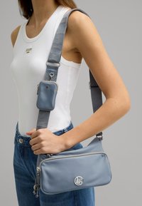 La borsa a tracolla grigia presenta un materiale liscio, hardware in argento, una tasca con zip e una tracolla regolabile a fantasia.