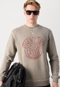 Grå hettegenser med et rundt rødt logo som har teksten "TWENTY TWO SCOTCH & SODA AMSTERDAM." Modell som holder en svart veske, iført svarte solbriller.