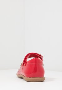 Zapato de charol rojo con correa trasera, punta redondeada y suela de goma marrón claro. Muestra detalles de diseño perforados en los lados.