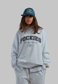 Szare, oversize'owe hoodie z napisem "POCKIES" oraz "Uniwersytet Marnych Osiągnięć". W połączeniu z jasnoszarymi spodniami dresowymi. Czapka w niebieskim kolorze z logo.