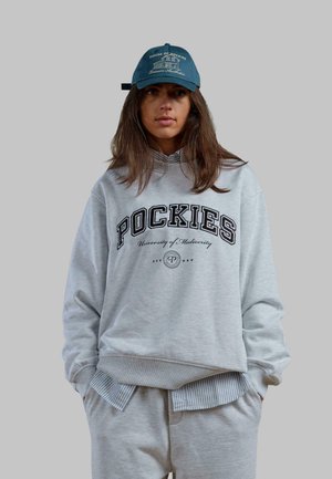 Felpa grigia over-sized con la scritta "POCKIES" e "Università della Mediocrità". Abbinata con pantaloni della tuta grigi chiari. Cappellino di colore blu con logo.