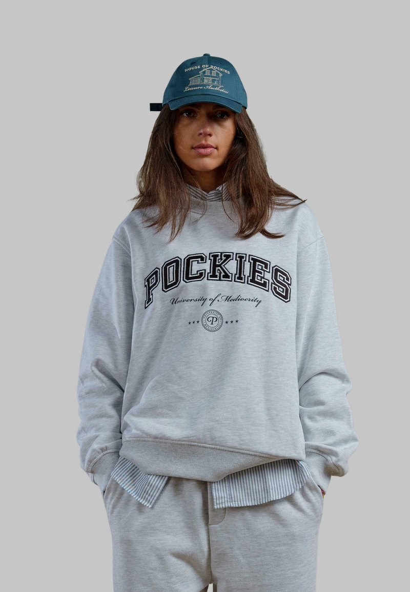 Szare, oversize'owe hoodie z napisem "POCKIES" oraz "Uniwersytet Marnych Osiągnięć". W połączeniu z jasnoszarymi spodniami dresowymi. Czapka w niebieskim kolorze z logo.