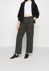 Giacca nera in velour con zip e cappuccio, top corto bianco, pantaloni cargo verdi a gamba larga con tasche e sneakers nere chunky.