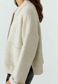 Beige gebreide cardigan met een kraag, verlaagde schouders en een ontspannen pasvorm. Heeft een textuurpatroon en twee voorzakken.