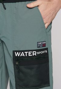 Vízisportos rövidnadrág világoszöld anyagból, fekete hálós zsebbel, cipzárral, fehér "WATER SPORTS" felirattal és márkás öltészel.