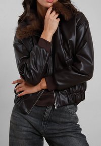 Giacca bomber in finta pelle marrone con collo di pelliccia, polsini a coste, orlo elastico e chiusura con zip; presenta una tasca laterale e una texture liscia.