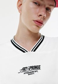 Ung person iført en hvid skjorte med sort stribet krave og teksten "ARTSPRINGS ATHLEISURE DPT. SINCE 91", samt en rød hat.