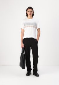 Weißes T-Shirt mit horizontalen grauen und pinken Streifen, kombiniert mit schwarzen Hosen. Das Model hält eine schwarze Tragetasche und trägt schwarze Schuhe.