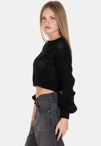 Maglione cropped nero in maglia con vestibilità ampia e maniche lunghe a sbuffo, che presenta un design aperto e texturizzato. Abbinato a jeans grigi a vita alta.