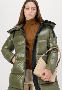 Olijfgroene pufferjas met een glanzende textuur en zwarte voering, die over een bruine gebreide coltrui is gestyled, gecombineerd met een beige handtas.