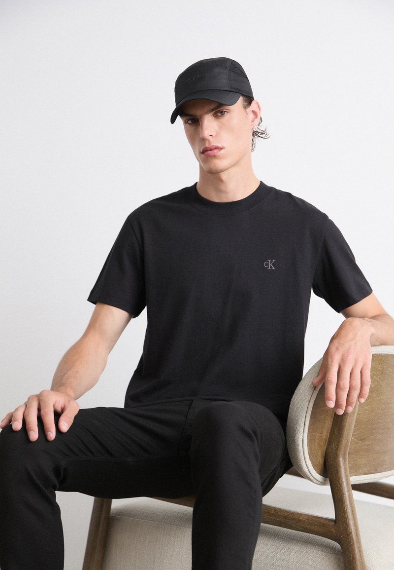 T-shirt in cotone nero a maniche corte con un piccolo logo grigio, abbinato a pantaloni neri e un berretto nero abbinato con una texture liscia.