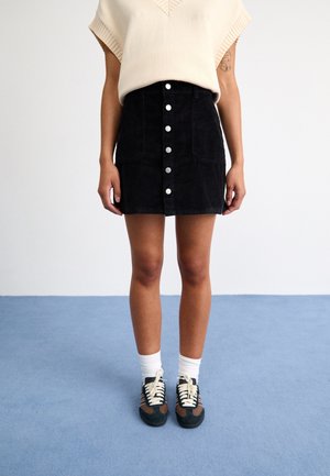 Personne portant un haut en tricot beige à manches courtes, une jupe en velours côtelé noire avec boutons à l'avant, des chaussettes blanches et des baskets noires et marron, debout sur un sol bleu.