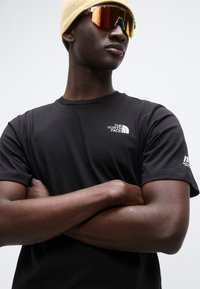 Μαύρο βαμβακερό κοντομάνικο μπλουζάκι, με το λογότυπο της The North Face σε λευκό στο στήθος και το μανίκι. Το μοντέλο φοράει κίτρινο σκουφάκι και ανακλαστικά γυαλιά ηλίου.
