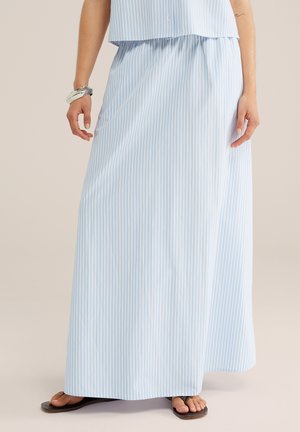 Femme portant une longue jupe rayée verticalement bleu clair et blanc avec des tongs noires et un bracelet en argent.