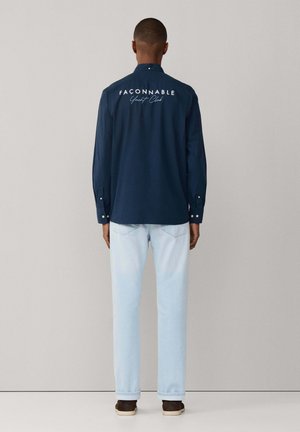 Hombre de espaldas con una camisa azul marino abotonada con el texto "Façonnable Yacht Club" en la espalda, pantalones vaqueros azul claro y zapatos oscuros.