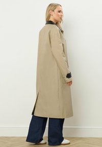 Lungo cappotto beige con colletto nero, fessure laterali e polsini con bottoni. Abbinato a jeans a gamba larga e sneakers bianche. Texture liscia.