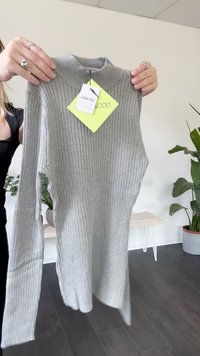 Person hält einen hellgrauen, gerippten Longsleeve-Pullover mit hohem Kragen und einem Zalando-Tag in einem Raum mit Pflanzen und einer Bank.