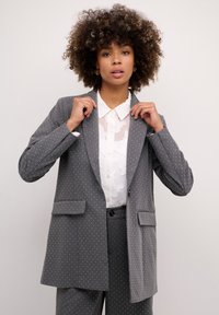 Pelēks uzplecu žakets ar peldošām punktiņām, divām kabat�ām, ar izgrieztiem lapelēm un brīvu piegriezumu, virs baltas teksturētas blūzes.