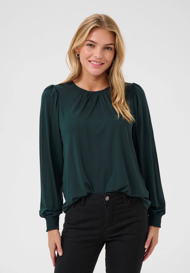 Blouse à manches longues vert foncé avec épaules froncées et poignets élastiques, associée à un jean noir ; texture lisse. Encolure ronde simple.