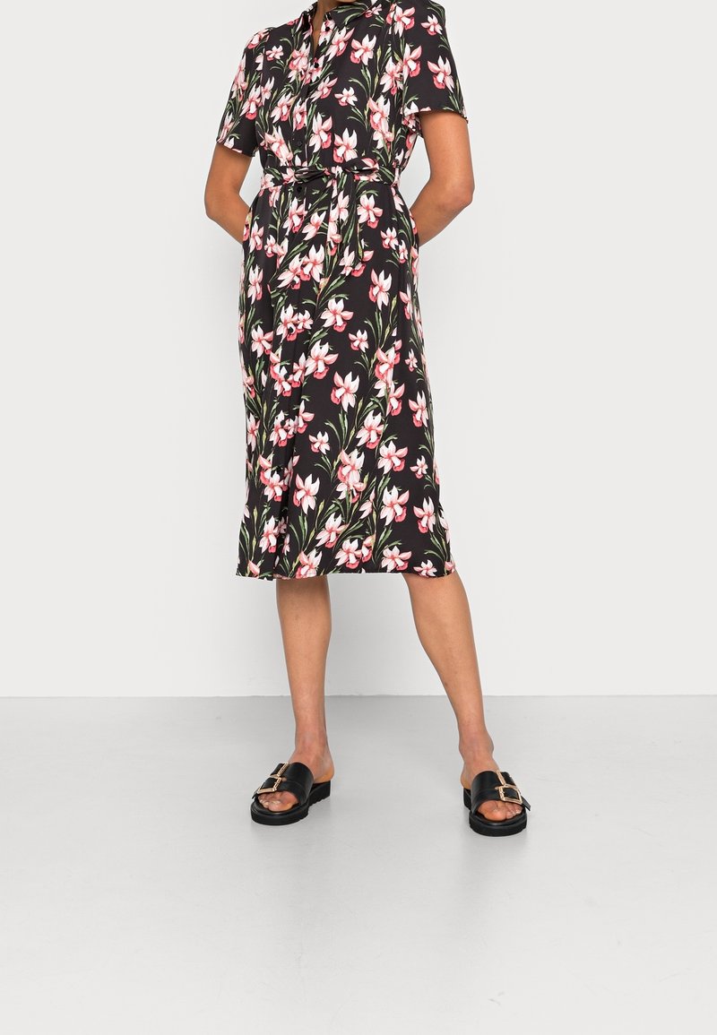 Femme portant une robe midi noire avec un imprimé floral rose et blanc, manches courtes et ceinture à la taille, associée à des sandales noires à boucle.