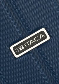 Material texturizado en color azul marino con una placa de logotipo rectangular plateada que lleva el texto "ITACA". La superficie presenta un patrón de puntos elevados y líneas diagonales.