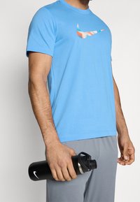 Sininen lyhythihainen Nike-t-paita, jossa on monivärinen logo. Henkilö pitää mustaa vesipulloa, jossa on hopeinen korkki. Jalassa on harmaat shortsit.