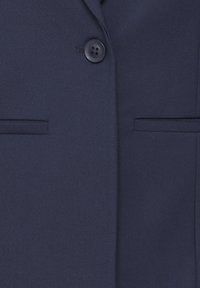 Gros plan sur un blazer bleu marine avec un seul bouton foncé et deux poches à fermeture passepoilée horizontales à l'avant.