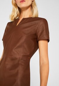 Brązowa skórzana sukienka z krótkimi rękawami, z dekoltem w kształcie łezki i szwami, które podkreślają dopasowany krój. Gładka faktura i minimalistyczny design.
