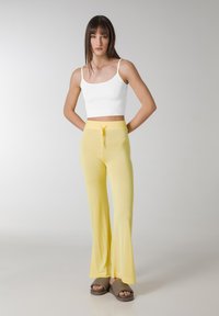 Pantalones acampanados de punto amarillos con cintura elástica y cordón, combinados con una camiseta sin mangas de croché blanca y ajustada. El conjunto presenta texturas suaves y un ajuste relajado.