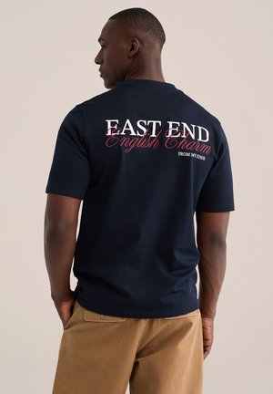 Mand iført en marineblå t-shirt med hvid og rød tekst ""EAST END English Charm FROM MY ENDS"" på bagsiden, kombineret med beige bukser.