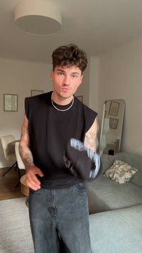 Top preto sem mangas, jeans de ganga, exibindo tatuagens nos braços. O fundo apresenta um sofá de cor clara e itens decorativos nas paredes.