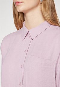 Woman wearing a light purple button-up shirt with a collar and chest pocket, showing her neck and lower face with silver hoop earrings.
