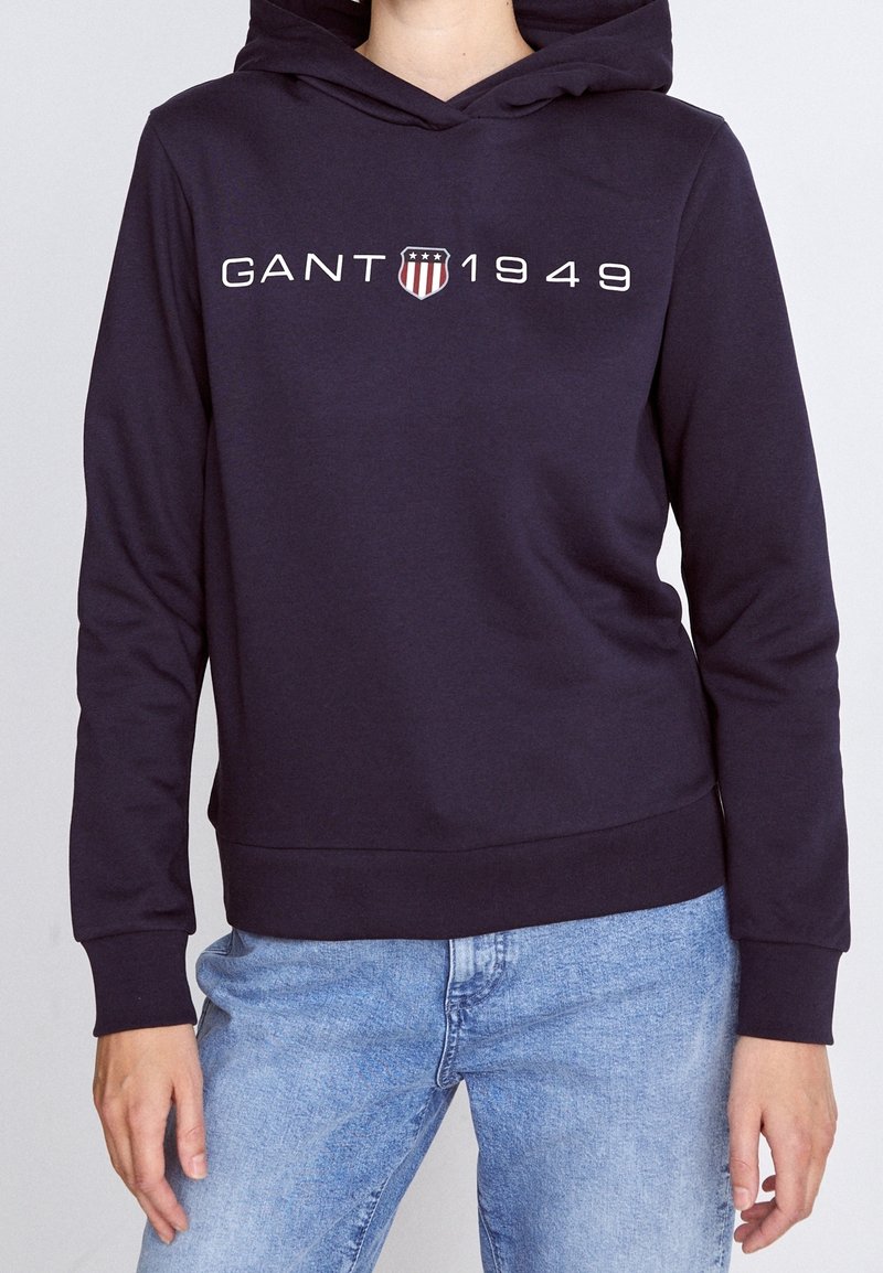 Femme portant un sweat à capuche bleu marine avec l'inscription "GANT 1949" et un logo de bouclier sur la poitrine, associée à un jean bleu clair.