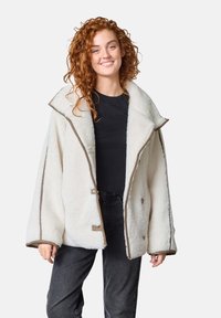 Överdimensionerad krämfärgad fleecejacka med en bred krage, bruna kanter, framslutande knappar och mjukt material, i kombination med en svart topp och mörka jeans.