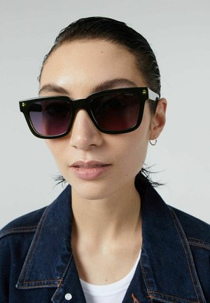 Lunettes de soleil carrées noires avec des verres teintés dégradés, dotées d'accents verts sur les branches et d'une finition brillante. Le modèle porte une veste en denim.