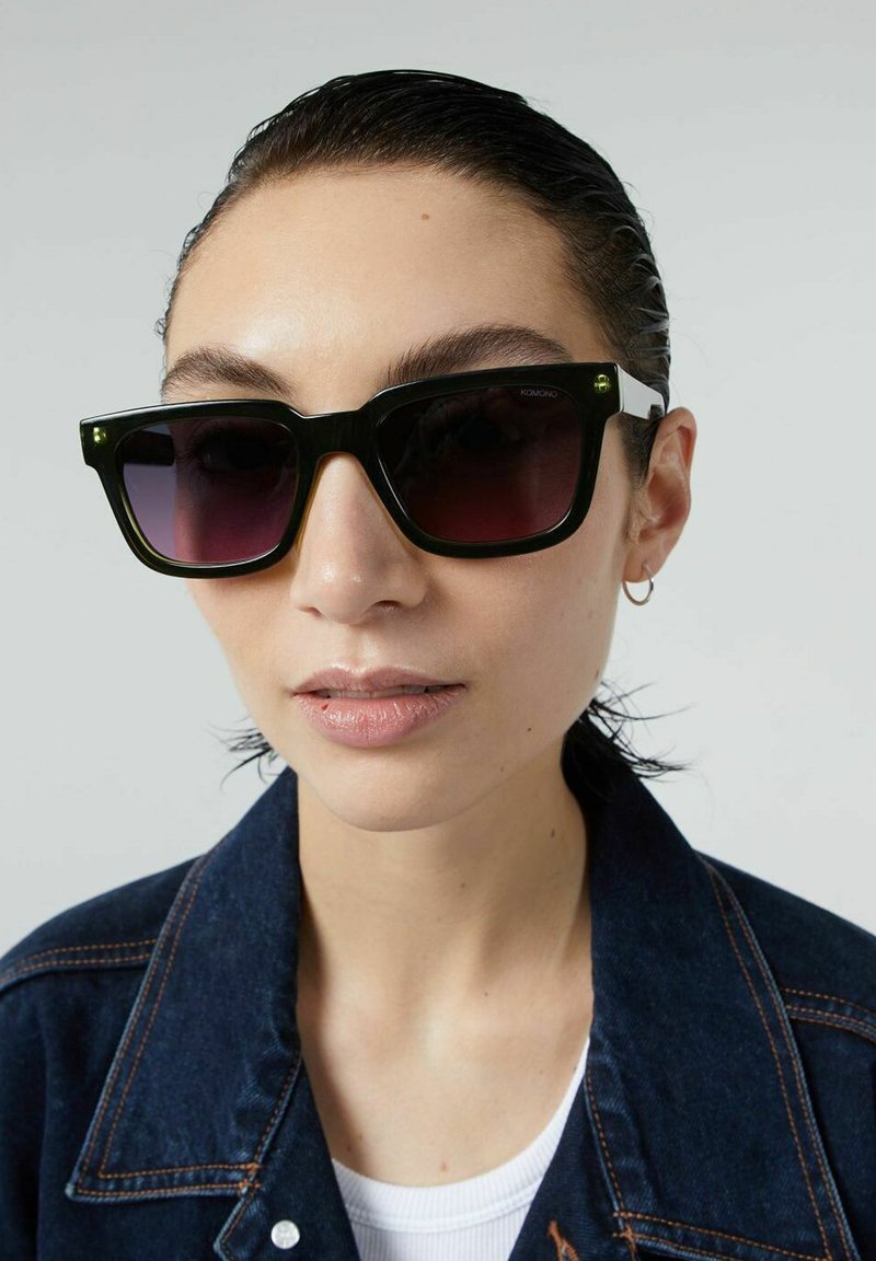 Zwarte vierkante zonnebril met een verloop van donkere lenzen, voorzien van groene accenten op de veren en een glanzende afwerking. Model draagt een denim jasje.