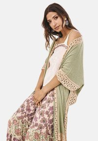 Chal verde de punto con borde de encaje; llevado sobre una blusa blanca y pantalones morados de volantes con estampado floral. Se ve la textura del tejido natural.