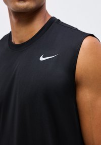 Haut du torse d’une personne portant un débardeur noir Nike avec un logo swoosh blanc sur la poitrine.
