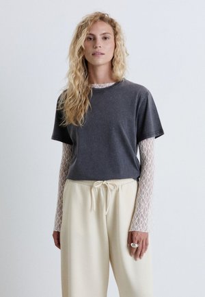 Donna bionda con capelli mossi che indossa una camicia scura a maniche corte sopra un top bianco in pizzo a maniche lunghe e pantaloni crema con coulisse.