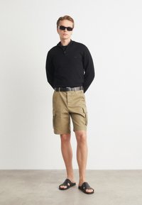 Bluză neagră cu mânecă lungă și fermoar, pantaloni scurți cargo khaki cu buzunare laterale, curea neagră, sandale negre și ochelari de soare închiși la culoare. Texturi netede și croială lejeră.