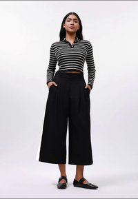 Nainen seisoo kädet taskuissa, päällään mustat leveät lyhyet housut, musta-valkoraidallinen pitkähihaista crop-top ja mustat kengät.