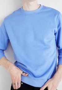 Sudadera azul claro hecha de una tela suave. Presenta un cuello redondo, puños y dobladillo de canalé. Llevada con una mano descansando en la cadera.