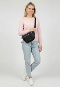 Žena v růžovém svetru, světlých modrých džínách, bílých teniskách a černé kabelce přes rameno stojící s překříženýma nohama.