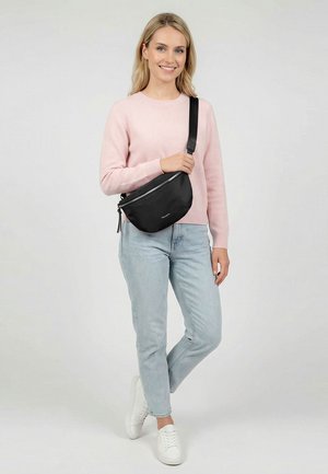 Frau trägt hellrosa Pullover, hellblaue Jeans, weiße Turnschuhe und schwarze Umhängetasche, steht mit übergeschlagenem Bein.