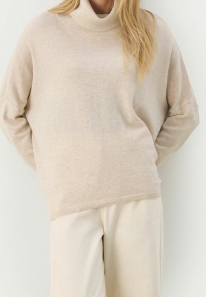 Femme portant un pull à col roulé beige et un pantalon couleur crème, debout les mains derrière le dos devant un fond uni.