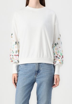Femme portant un sweat-shirt blanc à manches longues avec des motifs floraux multicolores sur les manches et un jean bleu clair, debout devant un fond uni.