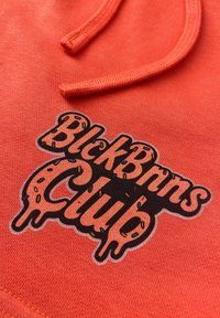 Oranje hoodie stof met een bedrukte logo waarop "BlickBros Club" staat, versierd met kleurrijke sprinkles op een zwarte achtergrond. Zichtbare decoratieve stiksels.